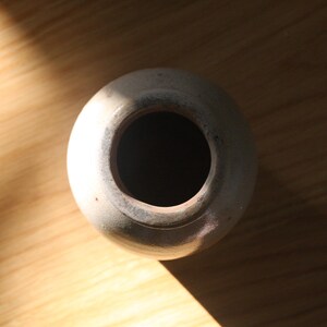 May include: A top-down view of a small, off-white ceramic vase with a wide opening. The vase sits on a light brown wooden surface, with a shadow cast across the wood. The vase has a simple, rounded shape.