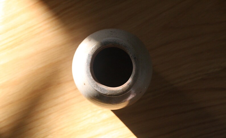 May include: A top-down view of a small, off-white ceramic vase with a wide opening. The vase sits on a light brown wooden surface, with a shadow cast across the wood. The vase has a simple, rounded shape.
