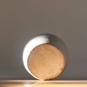 May include: A ceramic vase, viewed from the bottom, with a rounded shape and a light-colored, textured surface. The base is a lighter, unglazed color. The vase is set against a neutral background, with sunlight illuminating the object.