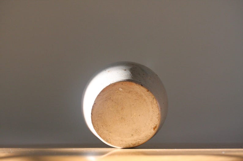 May include: A ceramic vase, viewed from the bottom, with a rounded shape and a light-colored, textured surface. The base is a lighter, unglazed color. The vase is set against a neutral background, with sunlight illuminating the object.
