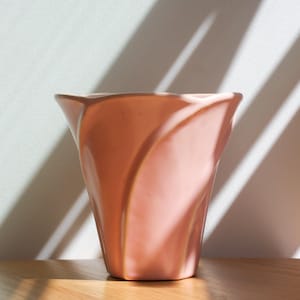 May include: A peach-coloured ceramic vase with a distinctive, flared form. The vase has a smooth, glossy finish and is set against a neutral background with shadows. The design is modern and elegant, suitable for displaying flowers or as a decorative piece.