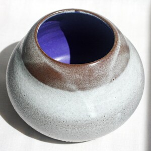 May include: A ceramic bowl with a rounded shape. The exterior is a speckled white and gray, with a band of brown near the top. The interior is a vibrant purple. The bowl is likely handmade.