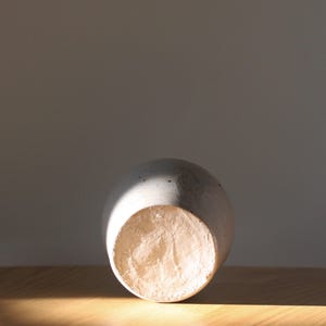 May include: A light gray ceramic vase with a rounded body and a textured, unglazed base. The vase is lying on its side on a wooden surface, with sunlight casting a shadow across it. The vase is a decorative item.