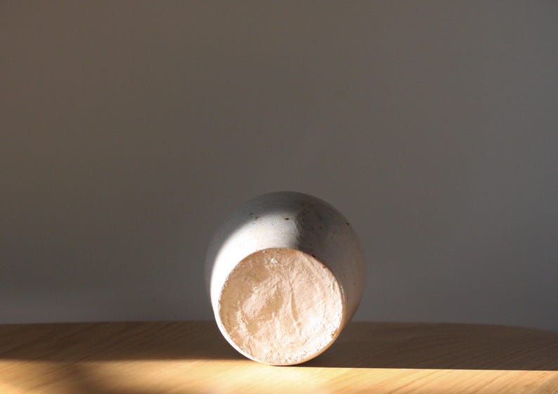 May include: A light gray ceramic vase with a rounded body and a textured, unglazed base. The vase is lying on its side on a wooden surface, with sunlight casting a shadow across it. The vase is a decorative item.