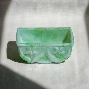 May include: A rectangular, green and white marbled glass planter. The planter has a curved front with two floral designs. The interior is a solid green color. The planter is sitting on a white surface.