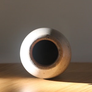 May include: A round, off-white ceramic vase with a dark interior. The vase has a wide opening and a slightly textured surface. The lighting highlights the vase's shape and the contrast between the light exterior and dark interior.