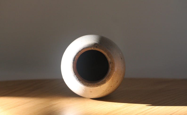 May include: A round, off-white ceramic vase with a dark interior. The vase has a wide opening and a slightly textured surface. The lighting highlights the vase's shape and the contrast between the light exterior and dark interior.