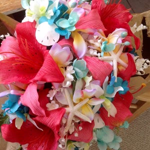 May include: A vibrant bridal bouquet featuring red, pink, and white flowers, accented with blue hydrangeas, starfish, and seashells. The bouquet is perfect for a beach wedding or tropical themed event.