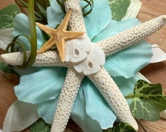 May include: A wedding bouquet with a starfish, sand dollars, and green leaves. The bouquet is wrapped in white satin ribbon and has a white flower on the side.
