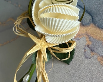 May include: A white seashell rose bouquet with a starfish and pearl accents. The bouquet is tied with brown raffia and green ribbon.