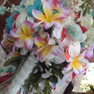 May include: A bouquet of artificial flowers in a clear glass vase. The bouquet features white, pink, yellow, and blue flowers with green leaves. The flowers are arranged in a circular shape and are accented with seashells.
