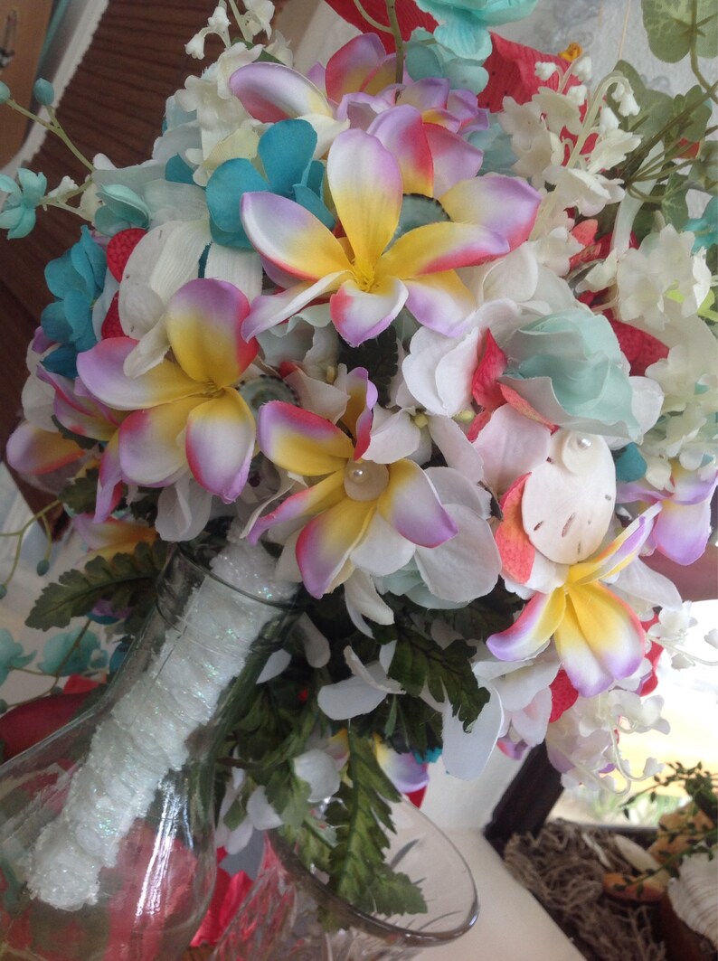 May include: A bouquet of artificial flowers in a clear glass vase. The bouquet features white, pink, yellow, and blue flowers with green leaves. The flowers are arranged in a circular shape and are accented with seashells.