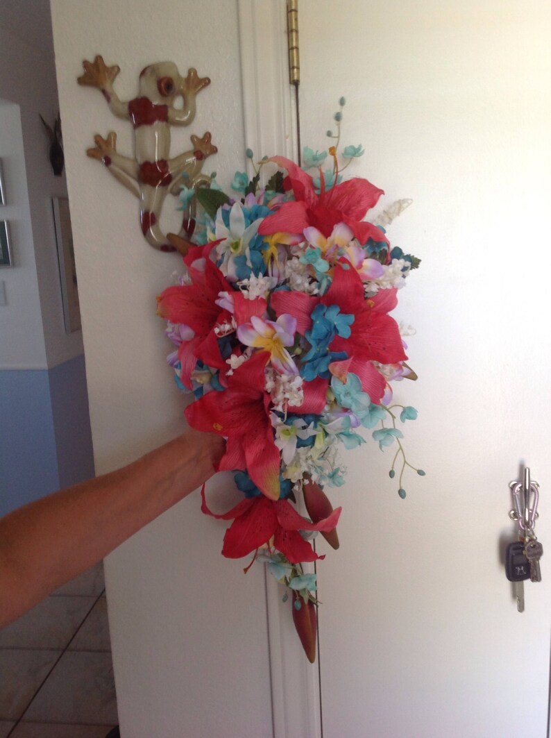 May include: A cascading bouquet of artificial flowers in shades of pink, red, blue, and white. The bouquet features large red lilies, smaller pink flowers, and blue hydrangeas.