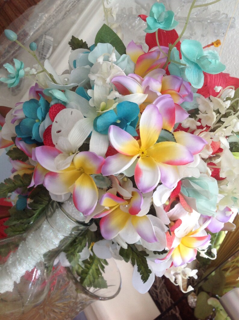May include: A bouquet of artificial flowers in shades of white, pink, yellow, blue, and red. The flowers are arranged in a clear glass vase with a white, sparkly ribbon.