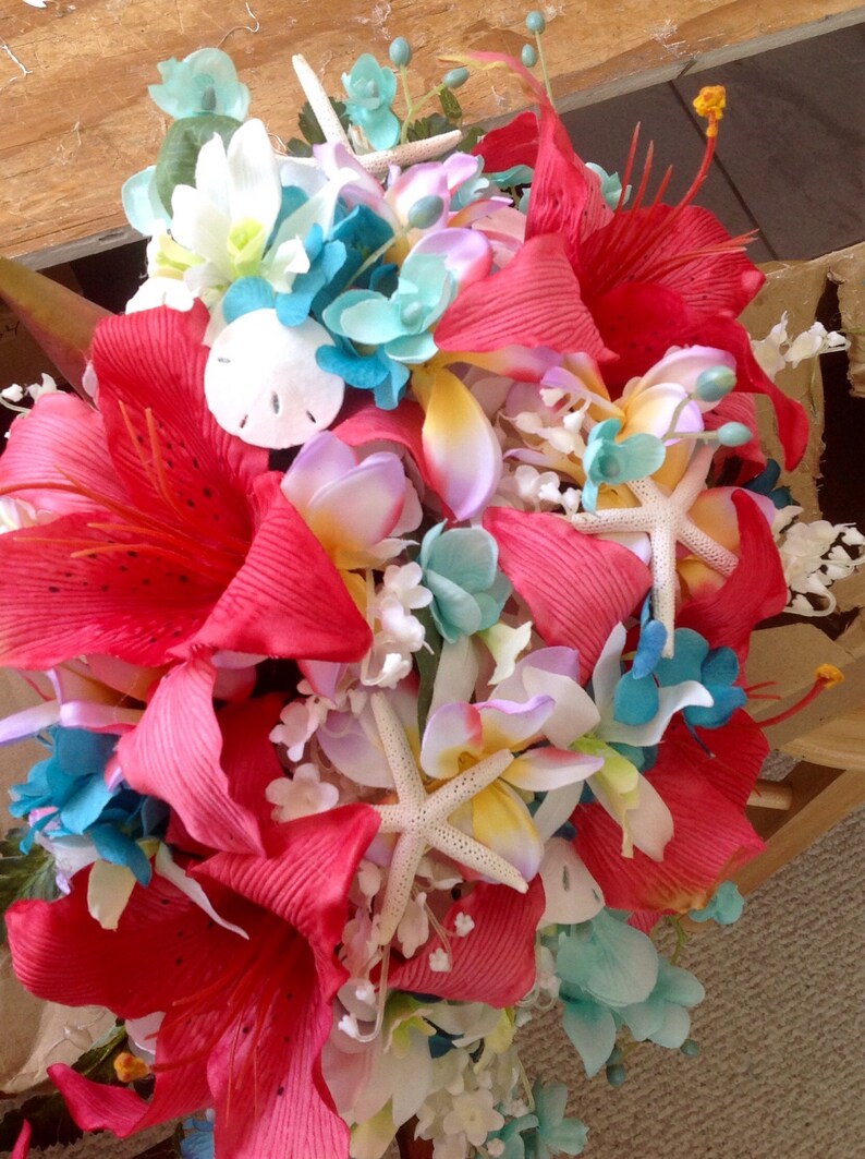 May include: A vibrant bridal bouquet featuring red, pink, and white flowers, accented with blue hydrangeas, starfish, and seashells. The bouquet is perfect for a beach wedding or tropical themed event.