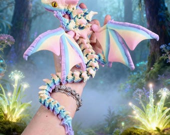 May include: A multi-colored articulated dragon figurine featuring a pastel gradient of pink, blue, and yellow. The dragon has large wings, a segmented tail, and prominent yellow eyes with black pupils, designed with a flexible, jointed structure.