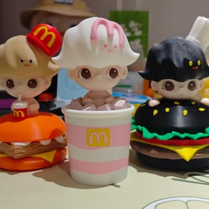May include: Three whimsical figurines, each styled after McDonald's food items. One is a burger, another a milkshake, and the third a burger with a chef's hat. Each figure has a unique design and color scheme, with the McDonald's logo visible.
