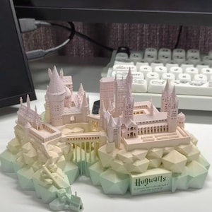 May include: A 3D printed model of Hogwarts School of Witchcraft and Wizardry featuring a pale pink castle structure atop a geometric, light green rocky base. The base includes a plaque with the text Hogwarts School of Witchcraft and Wizardry.