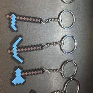 May include: Five pixelated keychains featuring light blue and dark brown block designs. Each keychain includes a metal ring and a short chain. The shapes represent various tools and a sword, all constructed with a distinct square grid pattern.