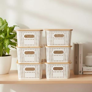 May include: Six white storage baskets with bamboo-colored lids are stacked on a wooden shelf. The baskets have a rectangular shape with a perforated design and a cutout handle. A potted plant and books are in the background.