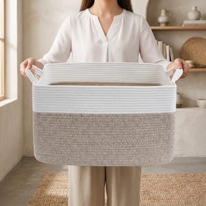 May include: A rectangular storage basket with a two-tone design. The top section is white, and the bottom section is a light brown. The basket has two white handles and is made of woven material. The basket is being held by a person.