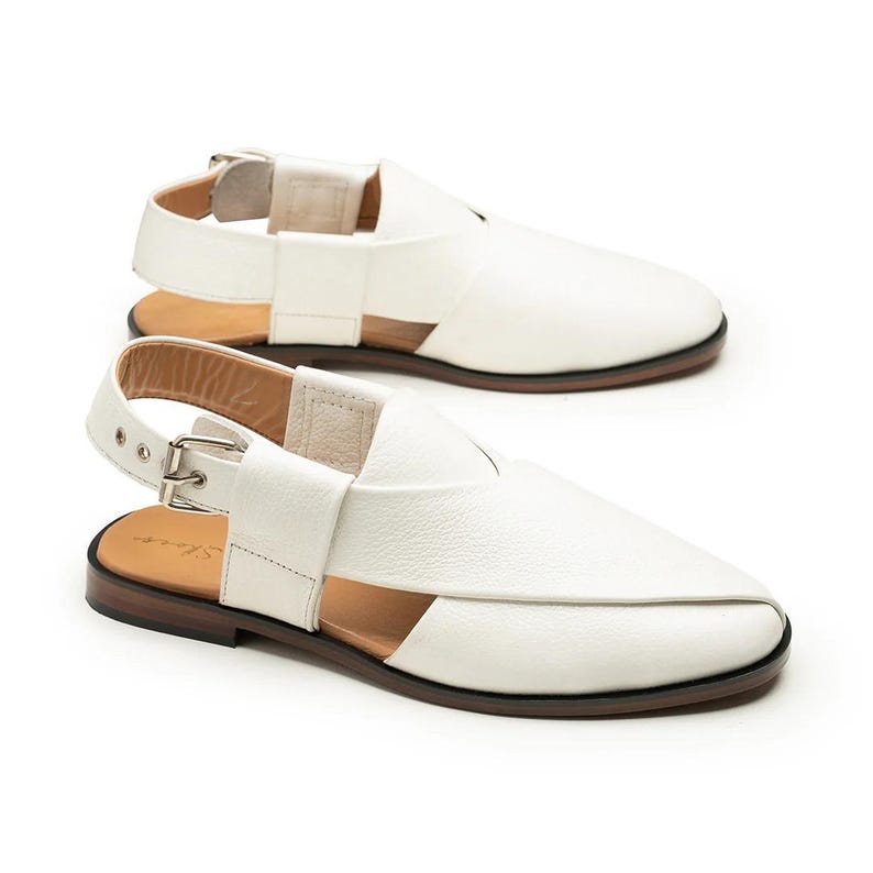 Puede incluir: Sandalias de cuero blanco con correa trasera y cierre de hebilla. Las sandalias tienen suela marr&oacute;n y un dise&ntilde;o recortado &uacute;nico. Los zapatos se muestran sobre un fondo blanco.