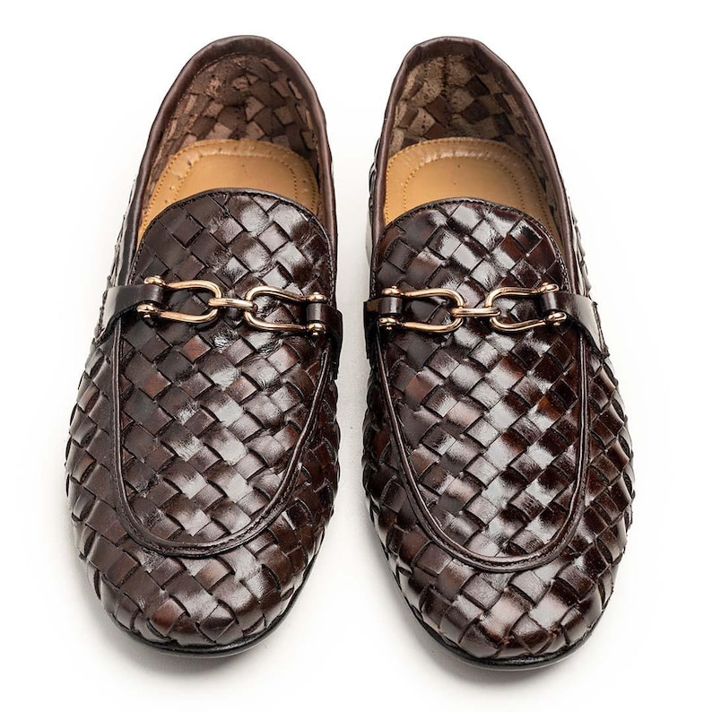 May include: A pair of dark brown woven leather loafers. The shoes have a gold-coloured chain detail across the top and a leather strap. The loafers have a rounded toe and a low heel. The shoes are displayed on a white background.