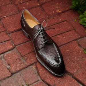 May include: A single brown leather dress shoe with a textured upper and a glossy toe cap. The shoe has dark laces and is set against a red brick background. The shoe is a classic style, suitable for formal occasions.