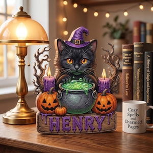 May include: A decorative wooden cutout featuring a black cat wearing a purple witch hat, positioned behind a green bubbling cauldron. Flanked by two jack-o'-lanterns and purple candles, the base displays the name HENRY in purple lettering.