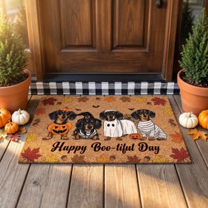 May include: A rectangular tan doormat featuring four dachshunds in Halloween costumes, including a pumpkin, a witch, a ghost, and a mummy. The mat is decorated with autumn leaves and the text Happy Boo-tiful Day in black script lettering.