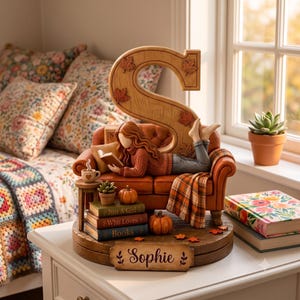 Puede incluir: Una figura decorativa con una letra S grande de madera, una persona leyendo en un sofá naranja y una pila de libros con el texto Just A Girl Who Loves Books. La base incluye una placa con el nombre Sophie y pequeñas calabazas naranjas.