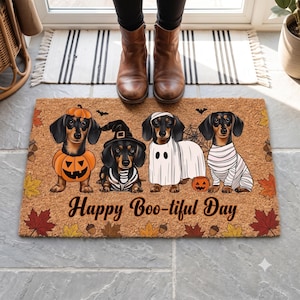 Puede incluir: Un felpudo rectangular de fibra de coco beige con cuatro perros salchicha disfrazados de Halloween: calabaza, bruja, fantasma y momia. La alfombra está decorada con hojas de otoño y bellotas, con el texto "Happy Boo-tiful Day" impreso en negro.