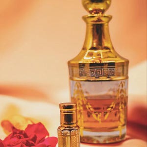 Op de afbeelding: Twee sierlijke, goudkleurige parfumflesjes met ingewikkelde ontwerpen. Eén is hoog met een afgeronde dop, de andere is klein en vierkant. Een rode bloem en saffraan op de voorgrond suggereren een geurthema.