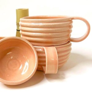 Może przedstawiać: Dwa kubki ceramiczne w kolorze jasnej brzoskwini z żebrowaną, poziomą fakturą na zewnątrz. Każdy kubek posiada szeroki, zakrzywiony uchwyt oraz gładkie, błyszczące wykończenie. Za kubkami widoczny jest fragment jasnobrązowego drewnianego narzędzia.