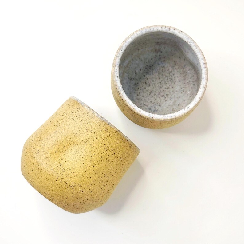 Może przedstawiać: Dwa ceramiczne kubki z nakrapianą ż&oacute;łtą powierzchnią zewnętrzną i jasnoszarym nakrapianym wnętrzem. Jeden kubek stoi pionowo, ukazując otw&oacute;r, podczas gdy drugi leży na boku, prezentując zaokrągloną podstawę i ż&oacute;łtą, teksturowaną powierzchnię.