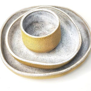 Może przedstawiać: Zestaw trzech ceramicznych naczyń składający się z dw&oacute;ch nieregularnych owalnych talerzy i małej miseczki. Każdy element ma teksturowaną musztardowo-ż&oacute;łtą powierzchnię zewnętrzną oraz nakrapiane biało-szare wnętrze z gładkim szkliwem.