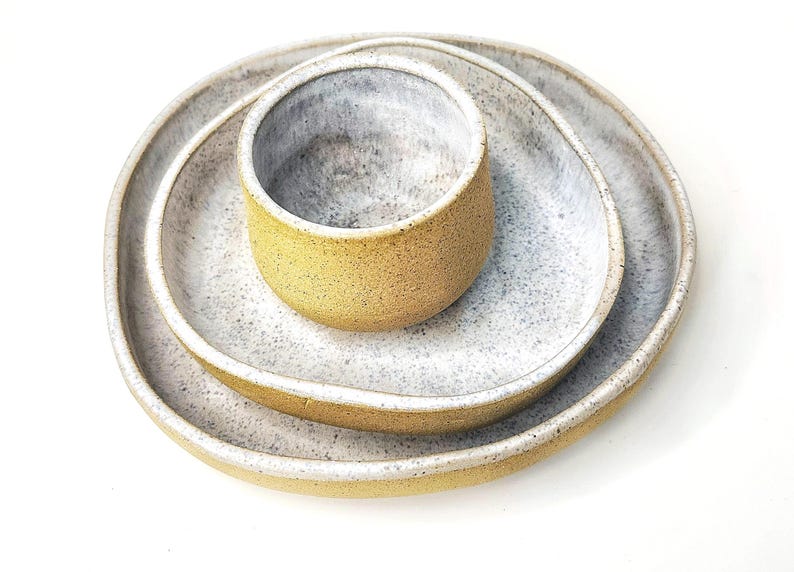 Może przedstawiać: Zestaw trzech ceramicznych naczyń składający się z dw&oacute;ch nieregularnych owalnych talerzy i małej miseczki. Każdy element ma teksturowaną musztardowo-ż&oacute;łtą powierzchnię zewnętrzną oraz nakrapiane biało-szare wnętrze z gładkim szkliwem.