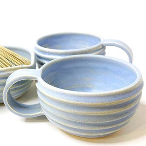 Może przedstawiać: Dwa jasnoniebieskie kubki ceramiczne z żebrowaną, poziomą fakturą na zewnątrz i jednym uchwytem. Trzecia ceramiczna miseczka zawierająca bambusową trzepaczkę jest częściowo widoczna w tle, pasując do jasnoniebieskiego koloru i żebrowanego wzoru.