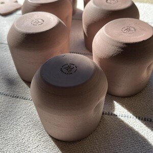 Może przedstawiać: Zestaw pięciu matowych, jasnor&oacute;żowych kubk&oacute;w ceramicznych ułożonych w grupę. Każdy kubek ma zaokrąglony, lekko nieregularny kształt oraz płaską podstawę z wytłoczonym napisem MIKKARES, wskazującym na markę i pochodzenie tych ceramicznych naczyń.