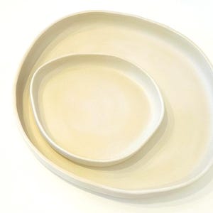 Może przedstawiać: Dwa kremowe ceramiczne talerze, jeden włożony w drugi. Talerze mają organiczny, nieregularny kształt z miękko zakrzywionymi krawędziami. Większy talerz jest w kolorze jasnokremowym, a mniejszy w nieco jaśniejszym odcieniu.
