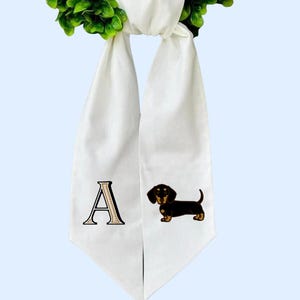 以下が含まれることがあります： 黒と茶色のダックスフント犬のイラストと文字「A」が描かれた白い卒業式用ストール。ストールは緑のリースから吊り下げられています。ストールは柔らかく流れるような生地で作られています。