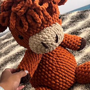 May include: A plush stuffed animal crafted from thick, textured orange yarn. The creature features a prominent, shaggy mane, a light beige muzzle, dark brown hooves, and small black eyes. The design mimics a highland cow with a soft, chunky knit.