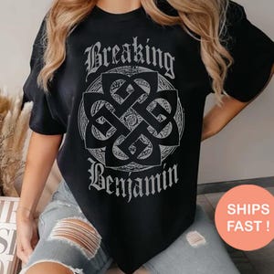May include: A black short-sleeved t-shirt featuring a white graphic print of a circular Celtic-style knot design centered on the chest. The text "Breaking" is printed in a gothic font above the knot, and "Benjamin" is printed below the knot.