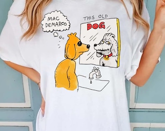 Op de afbeelding: Een wit t-shirt met een grafische illustratie van een gele hond die in een spiegel kijkt naar de reflectie van een oudere hond. De tekst "MAC DEMARCO" staat in een tekstwolk en "THIS OLD DOG" staat gedrukt boven de spiegel.