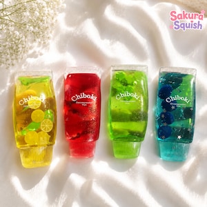 Könnte beinhalten: Vier transparente Quetschtuben mit der Aufschrift Chiboki Decompression toys. Die gelbe Tube enthält Zitronenscheiben, die rote Glitzer, die grüne Blattformen und die blaue Tube dunkelblaue Kugeln sowie silberne, sternförmige Pailletten.