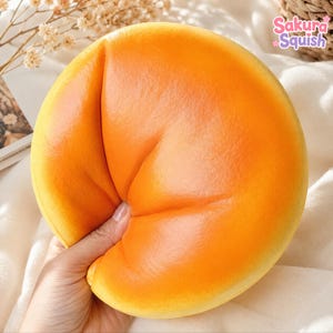 Può includere: Un giocattolo squishy rotondo di colore arancione con una consistenza superficiale liscia e compressa. L'oggetto è tenuto in mano e mostra una profonda rientranza dovuta alla pressione. Il testo Sakura Squish è visibile in alto a destra in rosa e viola.