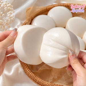 Può includere: Un giocattolo squishy bianco, liscio e rotondo, tirato per mostrare la sua consistenza flessibile. L'oggetto è tenuto sopra un cesto intrecciato contenente diversi squishy bianchi identici. Il testo Sakura Squish è visibile nell'angolo in alto a destra.