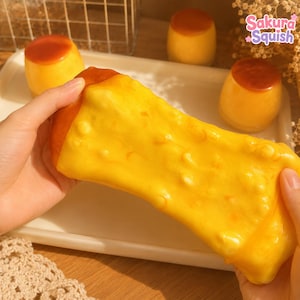 Può includere: Un giocattolo morbido rettangolare giallo con la parte superiore marrone, che ricorda un budino alla crema. Il giocattolo ha una superficie ruvida e irregolare e viene allungato. Sullo sfondo, tre giocattoli simili a budini su un vassoio bianco.