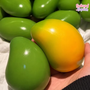 Può includere: Una collezione di giocattoli squishy lisci e arrotondati a forma di mango. Un giocattolo in primo piano è giallo brillante con una sfumatura verde, mentre gli altri sono verde tinta unita. Il testo Sakura Squish è visibile in alto a destra.