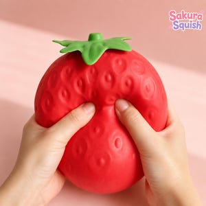 May include: A red strawberry-shaped squishy toy with a green stem and textured surface details. The toy is being compressed by two hands. The text Sakura Squish is visible in the upper right corner of the image in a stylised pink and white font.